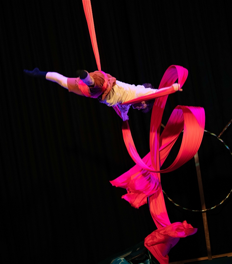 a big red silk is twisting in the air with an aerialist wearing earth tones is wrapped up in it. <br>the background is dark