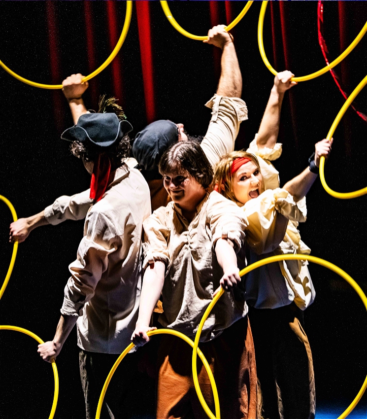 4 pirates are standing back to back with two looking towards the camera with cheeky smiles. they are holding hula hoops surrounding their bodies with yellow circles. <br>the background is dark .