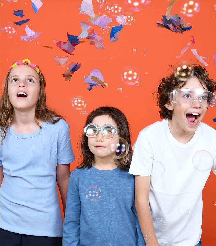 Three children wear goggles and stand against an orange backdrop. They all look thrilled. There are bubbles and pieces of coloured paper blowing around them.