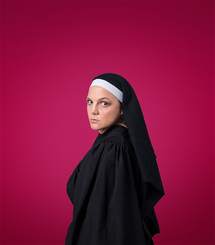 In front of a deep red background, a woman dressed as a nun looks over to the camera with a knowing look.
