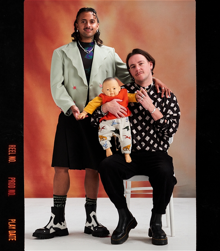 Krishna, a Brown trans person in their 30s, wears a green blazer and black skirt. Logan, their partner and a white trans person in their 30s, stands next to Krishna. Logan wears a black and white shirt and black pants, while holding a baby doll. Krishna and Logan are posed like they are in a family portrait, against an orange background.