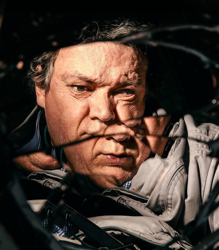 An older man stares into a broken mirror, his reflection is distorted by shards of glass.