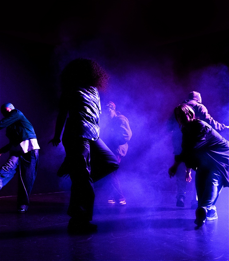 A hazy image of a group of people dancing around each in a dark room surrounded by smoke and deep blue and purple lights. The dancers are almost silhouetted and hold their own unique stances . The atmosphere is reminiscent of a club night.