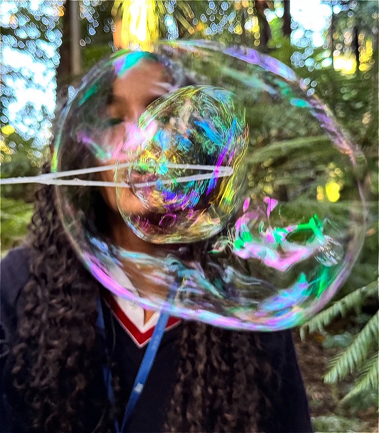 A teenage girl with long brown curly hair and a navy jumper blows a huge glistening rainbow bubble into the camera’s focus. She stands in a lush green forest surrounded by enormous green ferns.