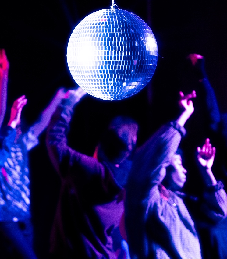 A hazy image and artfully blurred image of a group of people dancing energetically around each other with their hands upwards in the air. There is a shining disco ball, amongst deep blue, pink and purple lights in dark room. The camera is focused on the disco ball, located top middle of the image, while the dancers in the back are slightly blurred. The atmosphere is reminiscent of a club night.