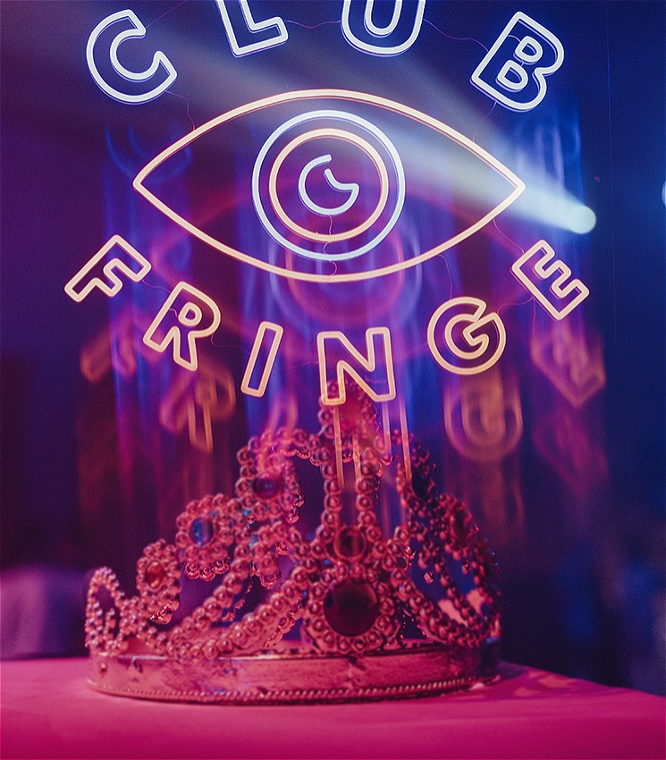 A hazy and artfully blurred image of a plastic crown lit in pink lighting. The image is overlayed with a neon logo which reads: CLUB FRINGE. In the middle of the two words is an the shape of an eye, with a crescent moon as the iris.