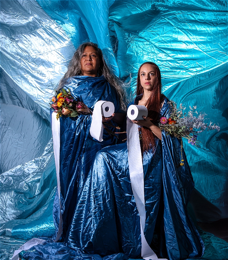 Two women stand side by side against a shiny blue fabric backdrop, wearing matching blue satin robes. Each holds a bouquet of flowers in one arm and a roll of toilet paper in the other, with the toilet paper unrolling downward. They both have stoic expressions.
