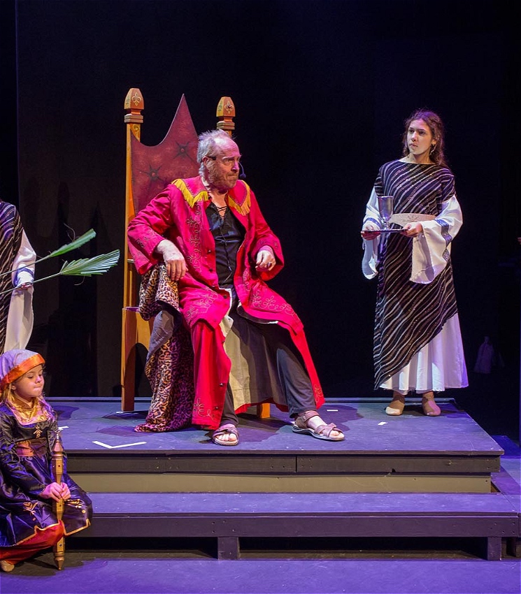 A live on-stage photo of a scene of actors against a black backdrop. An actor portraying Herod sits on a throne in his court. A woman stands to his right a a child kneels to his left.