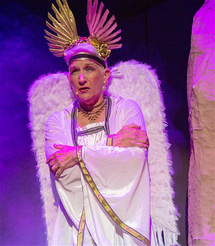 An actor wearing a head mic is dressed as an angel, They are all in white standing with their arms folded. They appear to be mid speech or song, and are staring out possibly to the audience who are out of frame. Their expression is focused and serious.