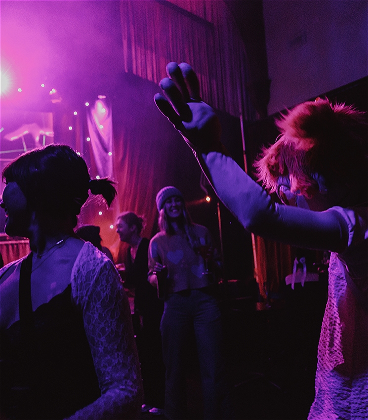 A group of people dancing in a dimly lit nightclub or performance space, illuminated by vivid pink and purple stage lighting. In the foreground, a person with bright red hair raises their arm while dancing, partially turned away from the camera. To the left, another person in a white lace top faces away, and in the background, others smile and move to the music. A stage with curtains and soft glowing lights is visible behind them, creating a lively, atmospheric scene.