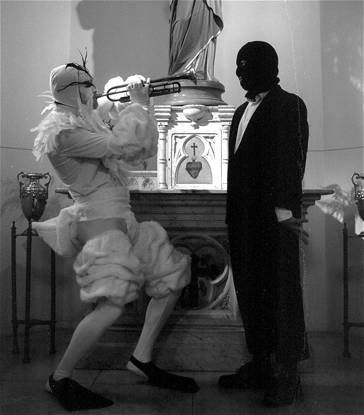 A black and white image of a trumpet player dressed as a white swan playing their instrument in the face of a person dressed in a dark suit with a black balaclava. They stand in front of a church statue of a figure with religious iconography that shows a cross and heart.