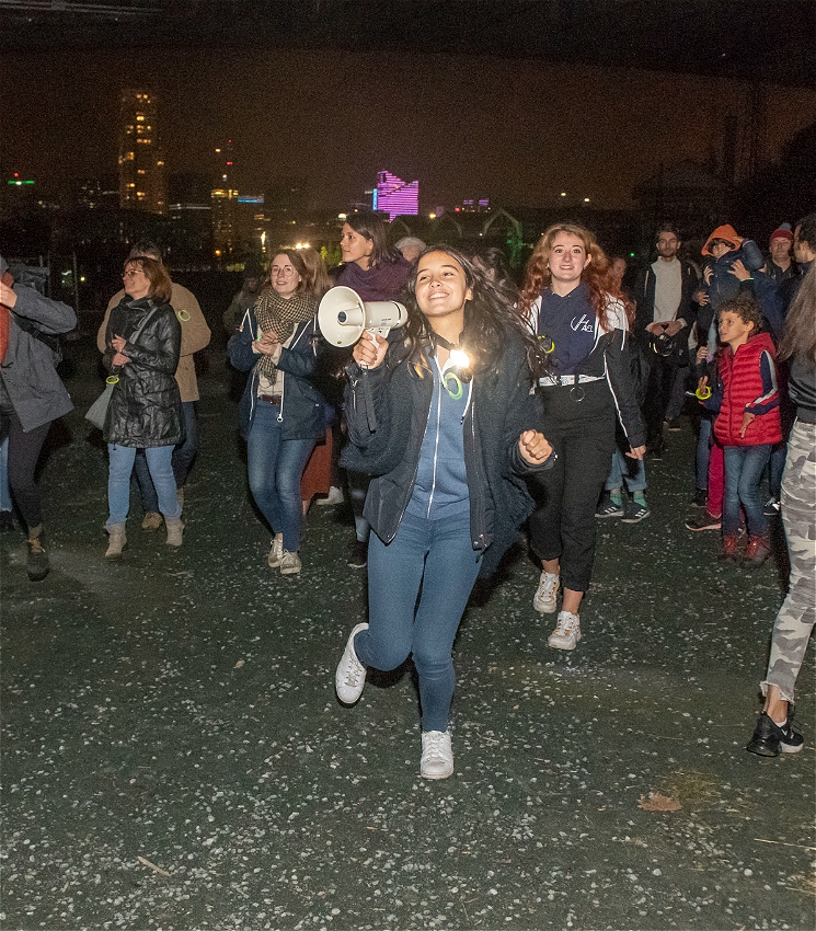A group of people of all ages walk through the streets at night. There is one person in denim jeans and a dark jacket who holds a loudspeaker.