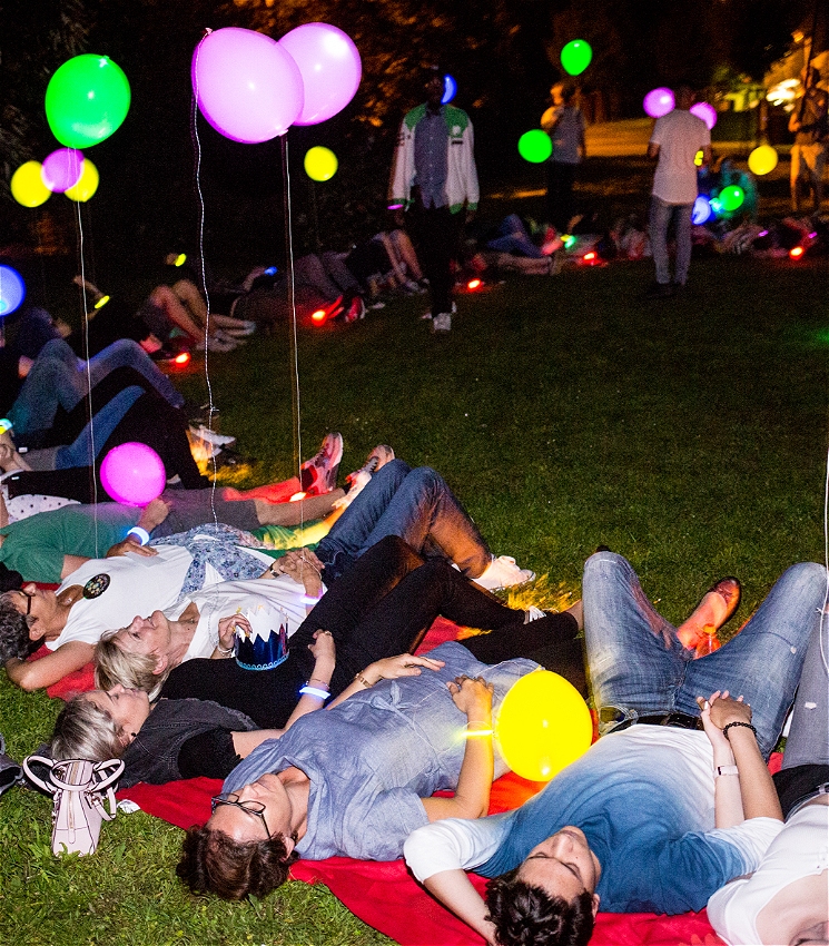 A group of people lying facing upwards on the grass in a circle at night. Some people hold colourful balloons on a string.