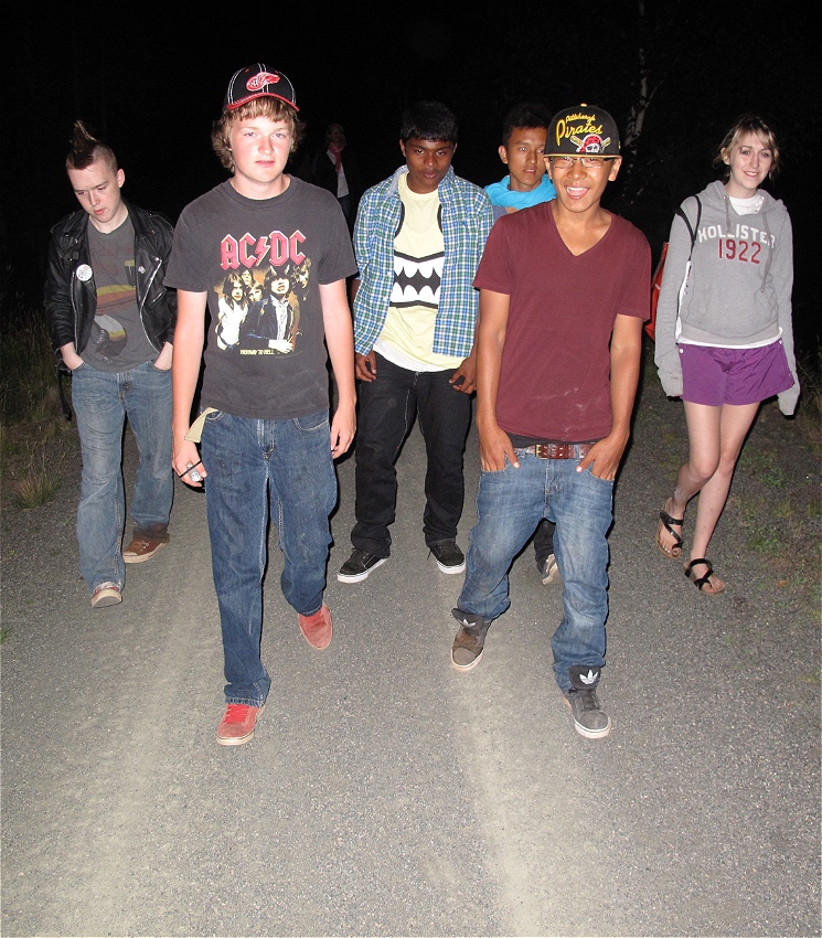 A group of six teenagers walk towards camera at night. Most of the teenagers wear jeans and t-shirts, some of them are smiling and some looking down at the ground.