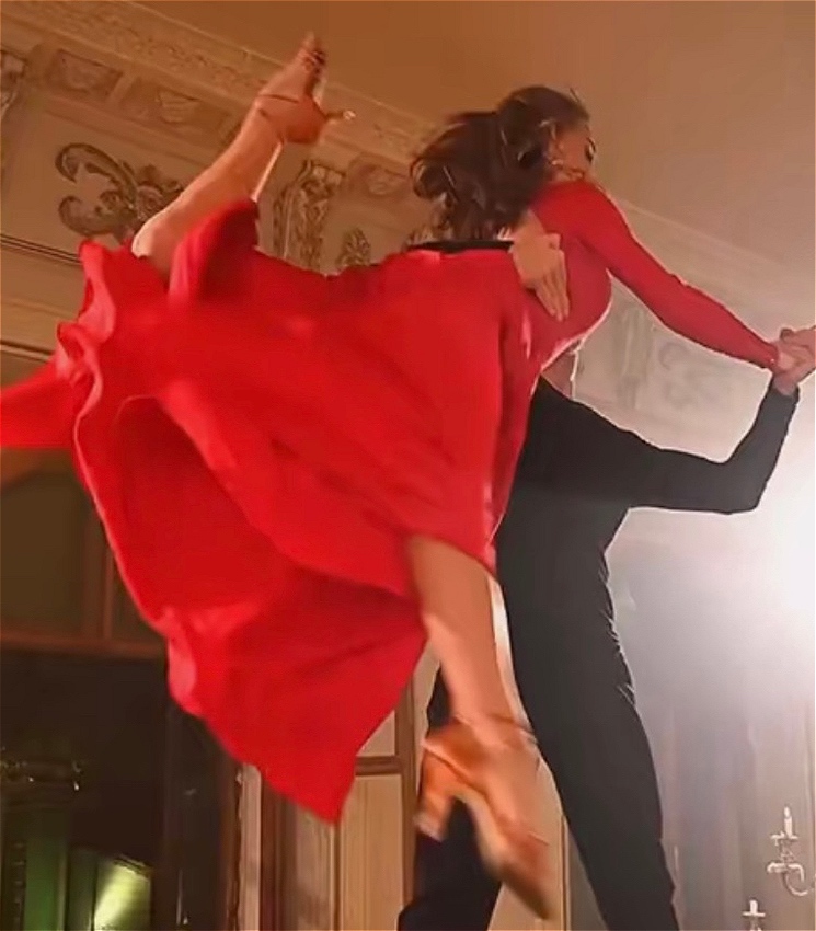 A woman in a red dress and a man in black dance. The woman is held up in the air by the man.