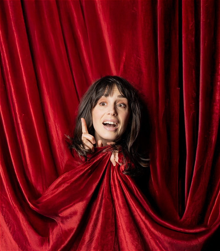 A woman with brown hair sticks her head out of a velvet red curtain, with an excited expression on her face. She points her finger in the air, and her other hand is clutching the red curtin under her face.
