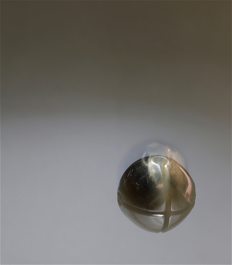 A close-up blurred shot that features a single, round bell, appearing to be made of a metallic or iridescent material. The bell is in the centre of the image against a soft, light grey background that darkens towards the top. The bell's smooth, reflective surface captures subtle light, highlighting its contours and suggesting its rounded shape. The camera angle is straightforward, placing the bell in the lower-middle right of the frame, with gentle lighting creating a smooth transition in the background.