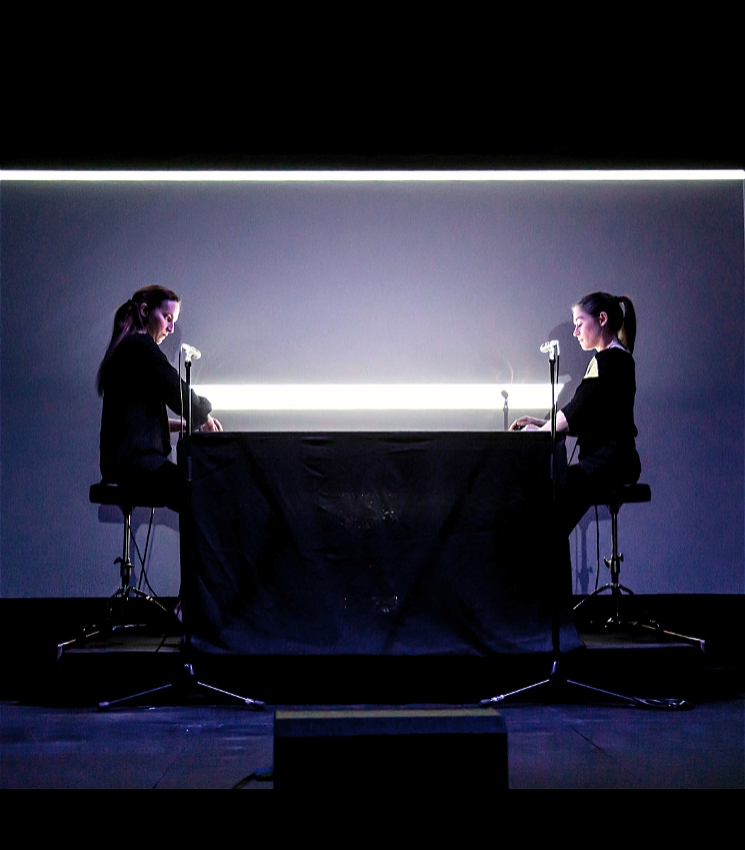 This dimly lit, wide shot shows two young women seated opposite each other at a long, black-draped table on a stage. Both women, facing inward, are dressed in dark clothing, with their hair pulled back, and each has a microphone positioned in front of them. Behind them, a bright, horizontal bar of light illuminates a large grey screen, creating a stark contrast with the dark foreground and highlighting their silhouettes.