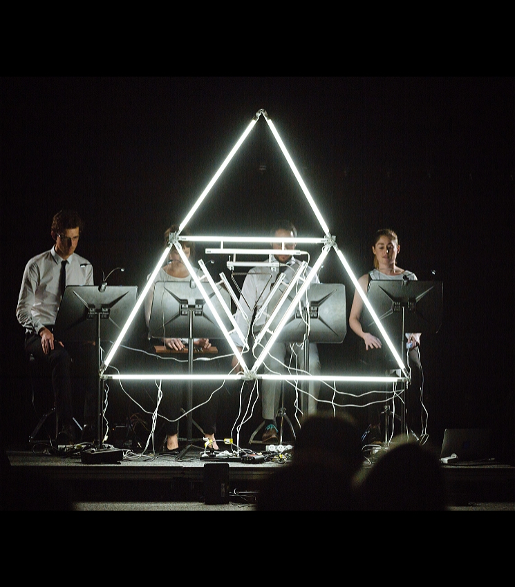 This wide, dimly lit shot features three people seated behind a large, illuminated triangular structure made of neon lights, creating a captivating focal point on a dark stage. From left to right, a man in a white shirt and tie, a woman in a grey top, and another woman in a striped top are each seated in front of their own music stands or screens, with various cables and equipment visible on the floor. The bright, geometric light sculpture contrasts sharply with the dark background, emphasizing the performers and their setup.
