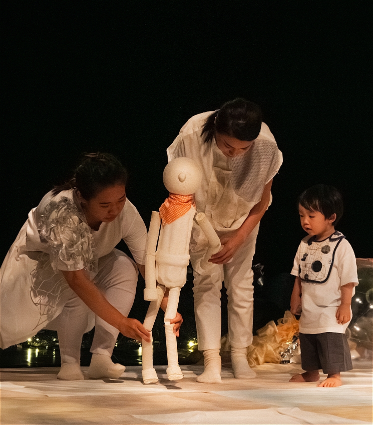 Two performers dressed in white outfits with silver detailing are moving a plain white puppet with an orange necktie. A toddler watches on, captivated by the puppet. The puppet is taller than the toddler.