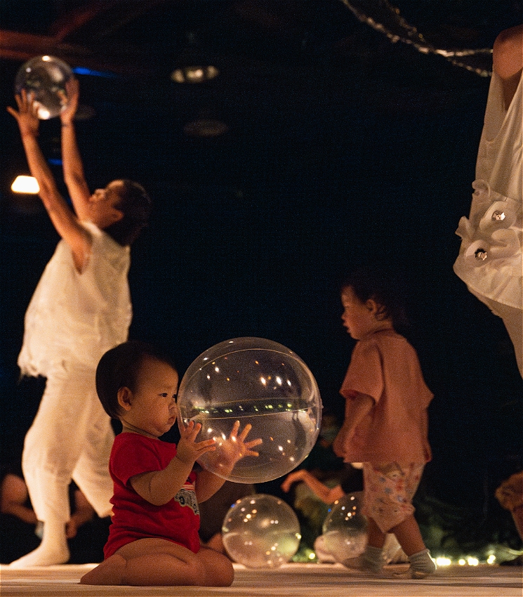 A toddler sits and holds an inflated clear ball, staring deep into its centre. In the background there are other toddlers, more balls, and performers dressed in white holding the balls up high.