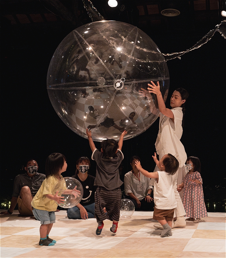 A performer dressed in white holds a large inflated clear sphere aloft. Reaching up for the ball are a collection of excited toddlers. In the background there parents and carers sitting down and watching on, looking amused.