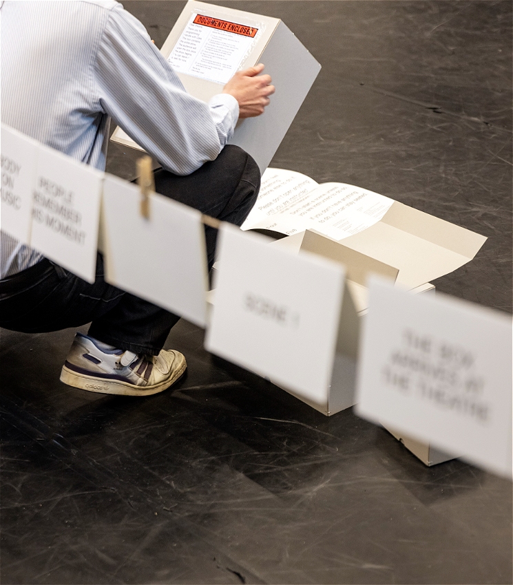 A person wears a striped shirt, jeans and sneakers and faces away from the camera. They are holding the lid of a box and reading instructions on a label, the instruction text is unable to be read. The person is positioned in front of a string which has printed cards attached to it with pegs, the text on the printed cards is out of focus.