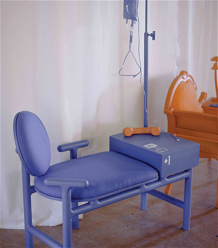 A stylised chair installation in soft blue tones, resembling a medical recliner with playful, rounded design features. An IV stand holds a matching blue fluid bag, and a retro orange phone handset rests on a side unit marked with a "+1" symbol and a thumbs-up icon. The scene is set against a white curtain backdrop with another quirky orange chair visible in the background.