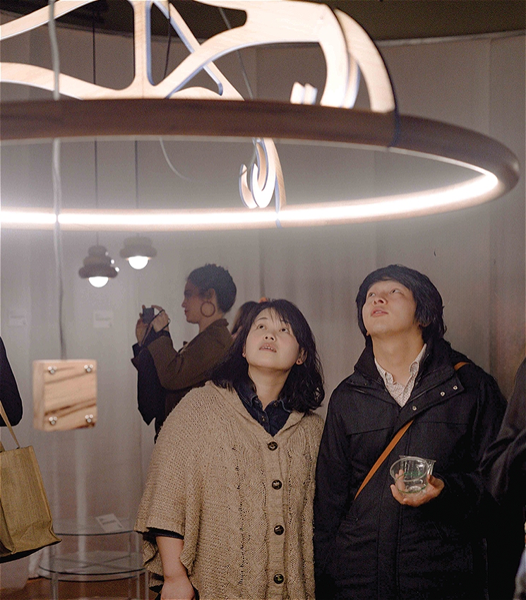 Two people stand closely together in a gallery space, gazing up in awe at a large, sculptural circular light fixture suspended above them. The installation is made of curved wooden elements and emits a soft glow. Other visitors are visible in the background, including one taking a photo, all surrounded by contemporary design objects displayed in a softly lit room.