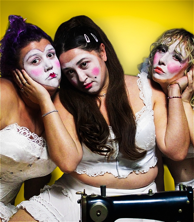 Three women sit close to one another. Their faces are painted white in the shape of a heart. Their cheeks are rouged and their lips lined red. They are sitting in front of a sewing machine and all have perplexed and slightly sad expressions. The middle woman cups the two women's face either side of her while they all look down the camera lens. One woman has purple hair and is wearing a white corset. The centred woman has brown hair and the woman to her right has blonde hair. All three are wearing lacey white garments.