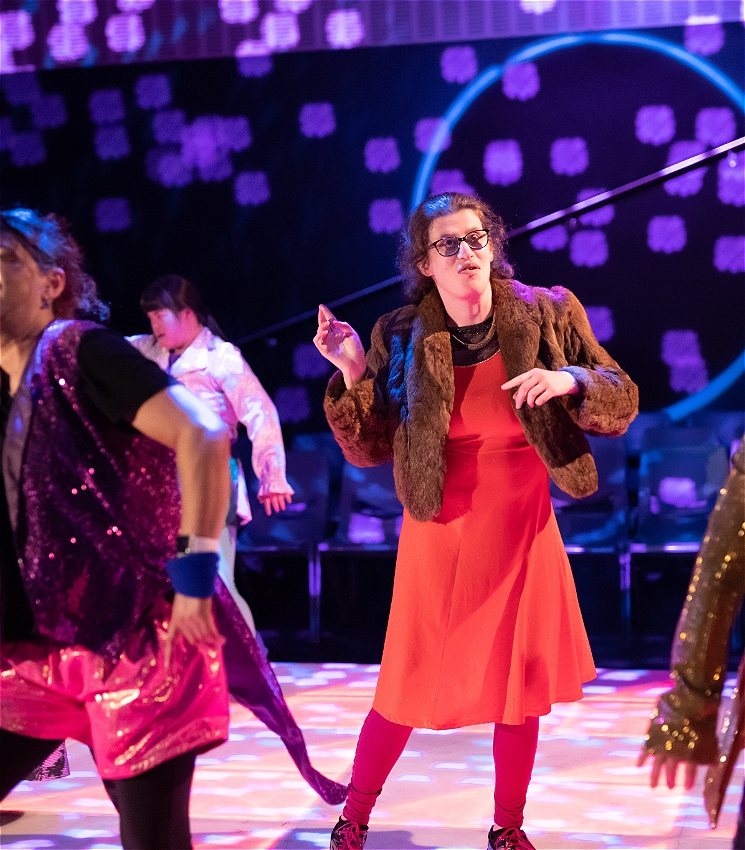 A group of performers dance at a disco. A dancer wearing a red dress and fur coat does a dance move with their arms raised up.