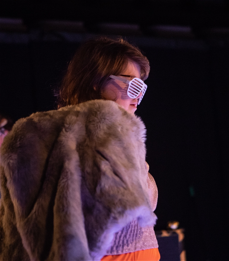 A person with sunglasses poses on the dancefloor with a fur coat around their shoulders. They are viewed in profile.