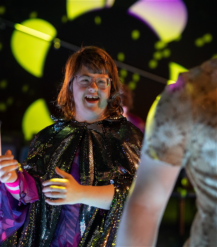 A dancer wearing white rimmed glasses and a sparkly cape, smiles on the dance floor.