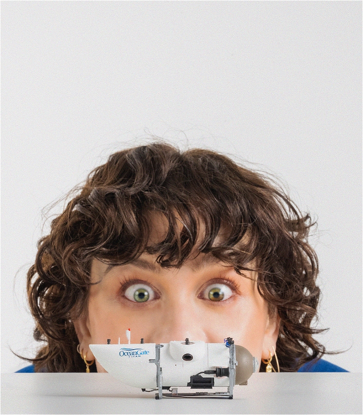 In the foreground, a small model of a deep-sea submersible rests on a white ledge - the words “OceanGate Titan” are just visible on its body. In the background, a young, white woman with curly brown hair is peeking up to look at it, her eyes lit up in excitement.