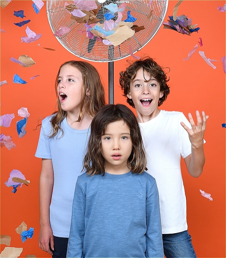 Three children stand against an orange backdrop and they look excited. A fan blows small pieces of coloured paper through the air around them.