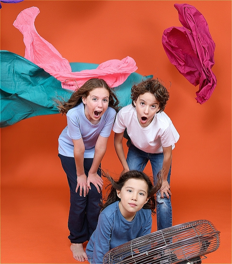 Three children stand against an orange backdrop. There is a fan in front of them that blows colourful pieces of fabric around their heads.