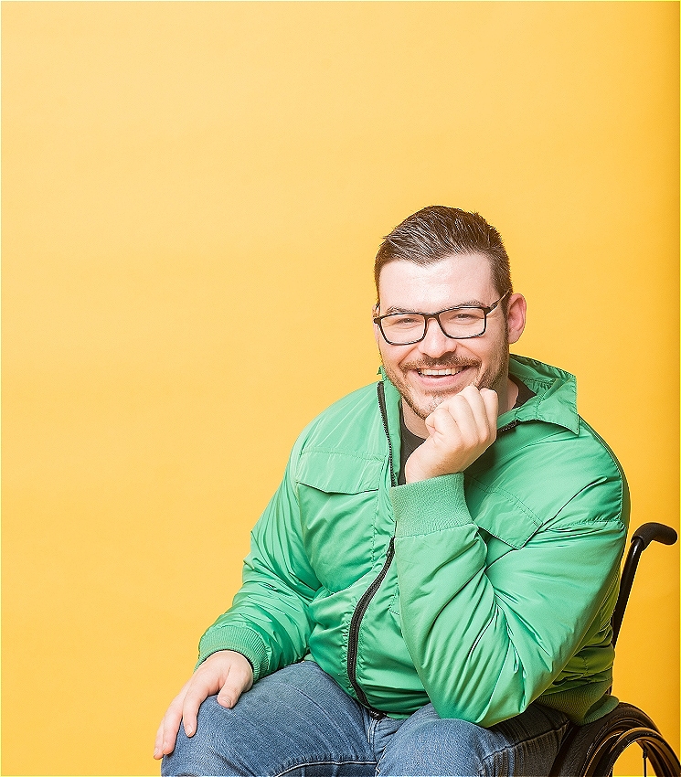 Oliver is sitting in his manual wheelchair in front of a yellow back drop. He is leaning forward in a relaxed position with his chin resting on his hand with a big grin on his on his face and an overall joyful vibe. <br><br>Oliver is wearing dark rimmed glasses, has short, dark hair with a well trimmed facial hair. He is wearing blue denim jeans and bright green bomber jacket