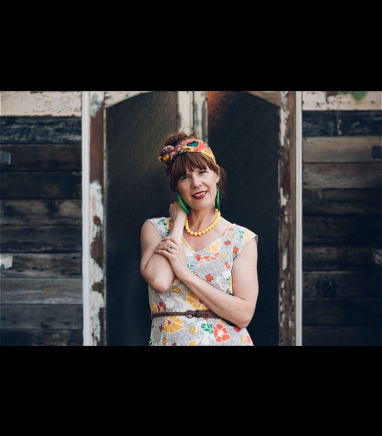 A woman leans against a rustic doorway with one arm crossed smiling at the camera. She wears green oversized earrings and yellow beads and a headscarf.