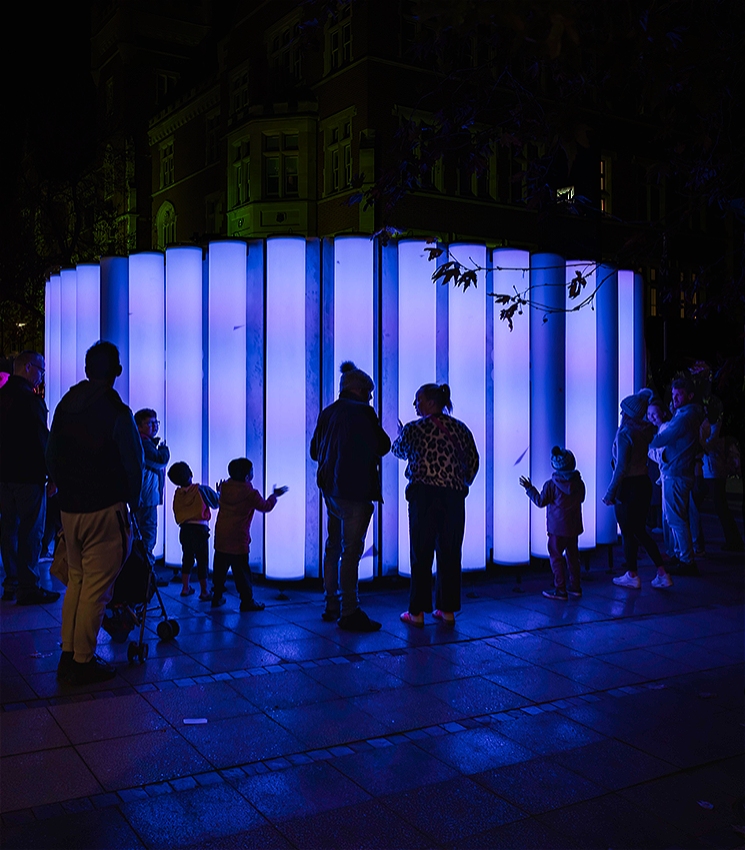 A crowd of people stands in front of the VOICES installation at night time. It is a vertical wall comprising blue, circular glowing light tubes.