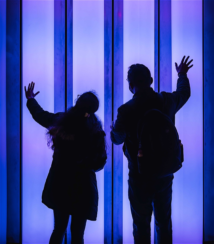 Silhouettes of two people standing in front of a glowing, vertical light installation with blue and purple hues. One person on the left has their hand raised to the lights, while the other on the right also reaches out with an open hand, both wearing coats and backpacks.