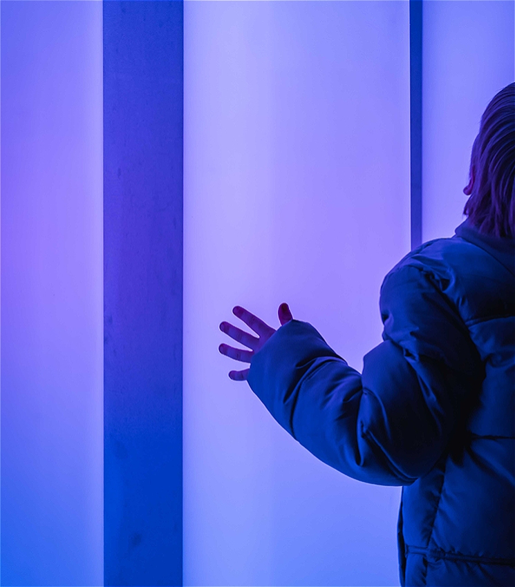 A person wearing a puffy jacket reaches out with one hand toward a glowing vertical light installation with soft blue and purple tones. Only the right side of their body is visible, highlighting their extended hand against the illuminated background.