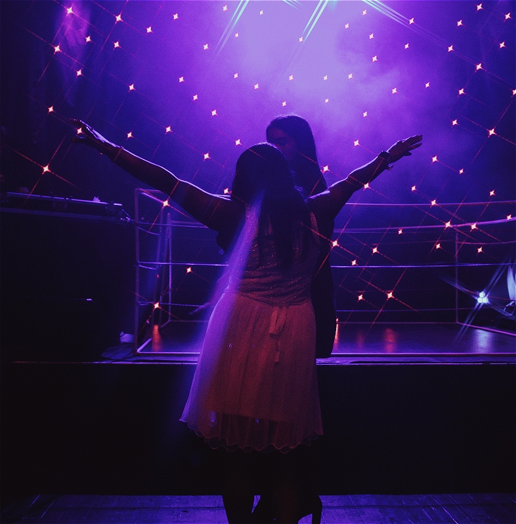 Two people dance under purple stage lights and a backdrop of star-like points of light. One person in front wears a light-colored dress and has their arms outstretched, while the other stands close behind them, partially obscured by haze and the sparkling lights.     Ask ChatGPT