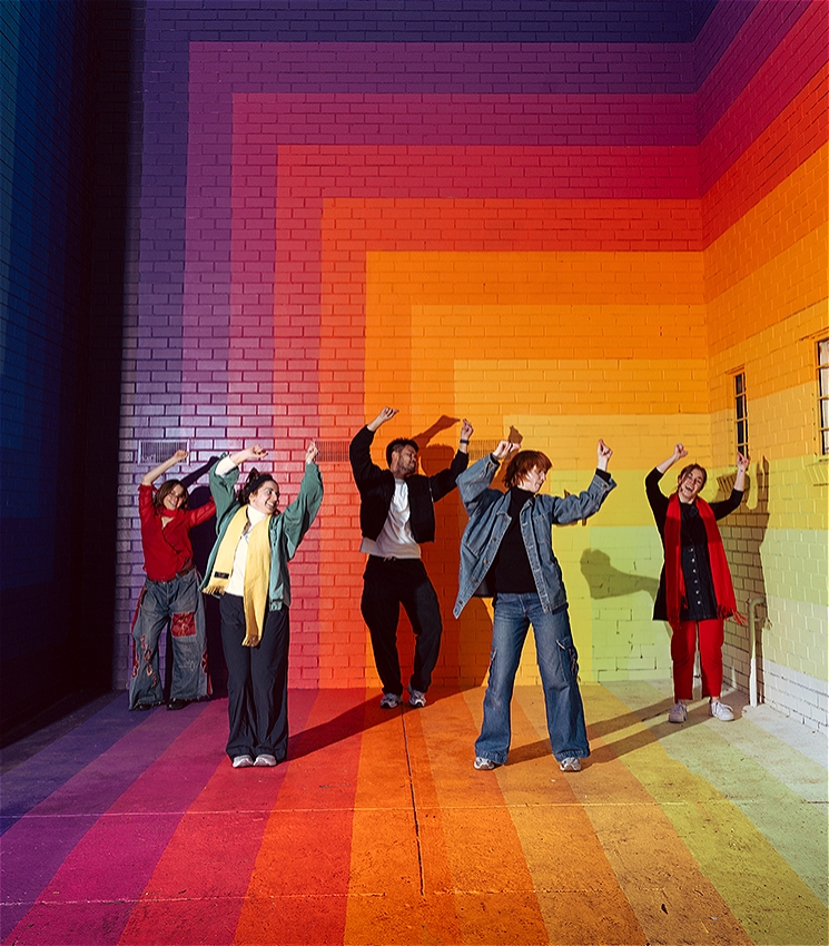 There are five dancers smiling and performing a dance move with their arms in the air. All of the dancers are in front of a brightly coloured painted rainbow mural by Drez.