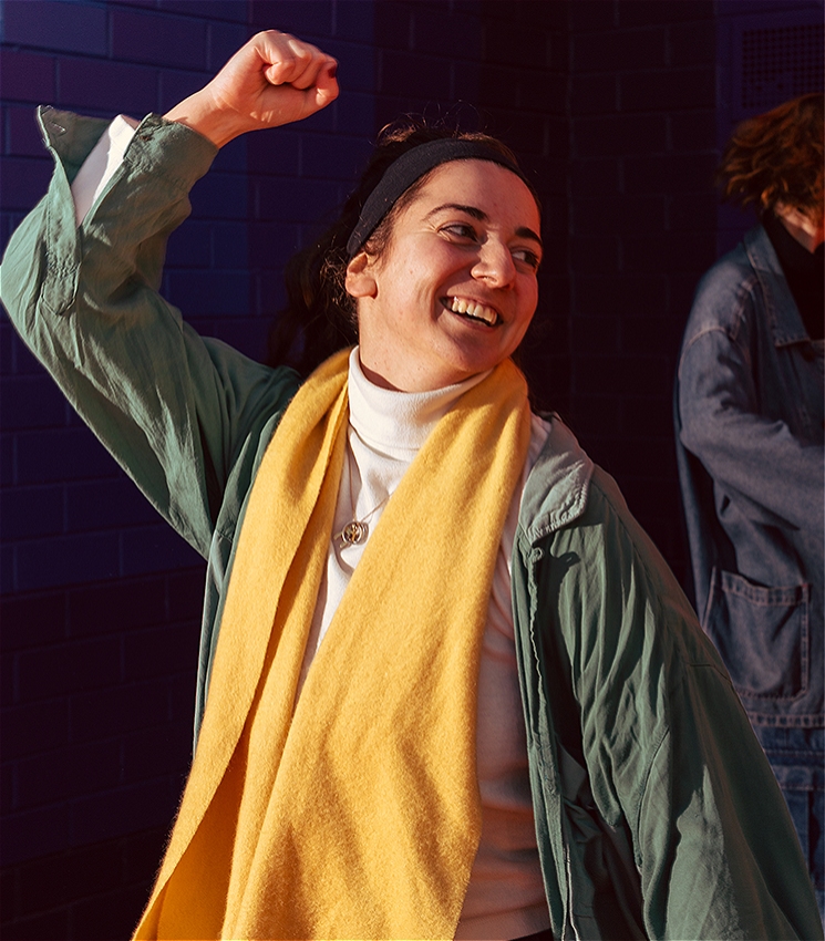 A dancer wearing a white top, a green jacket and a yellow scarf, smiles while stretching one arm above their head. There is a dancer wearing denim in the background.