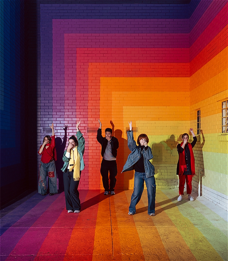 There are five dancers smiling and performing a dance move that involves one hand pinching their nose and one arm stretched above their head. All of the dancers are in front of a brightly coloured painted rainbow mural by Drez.