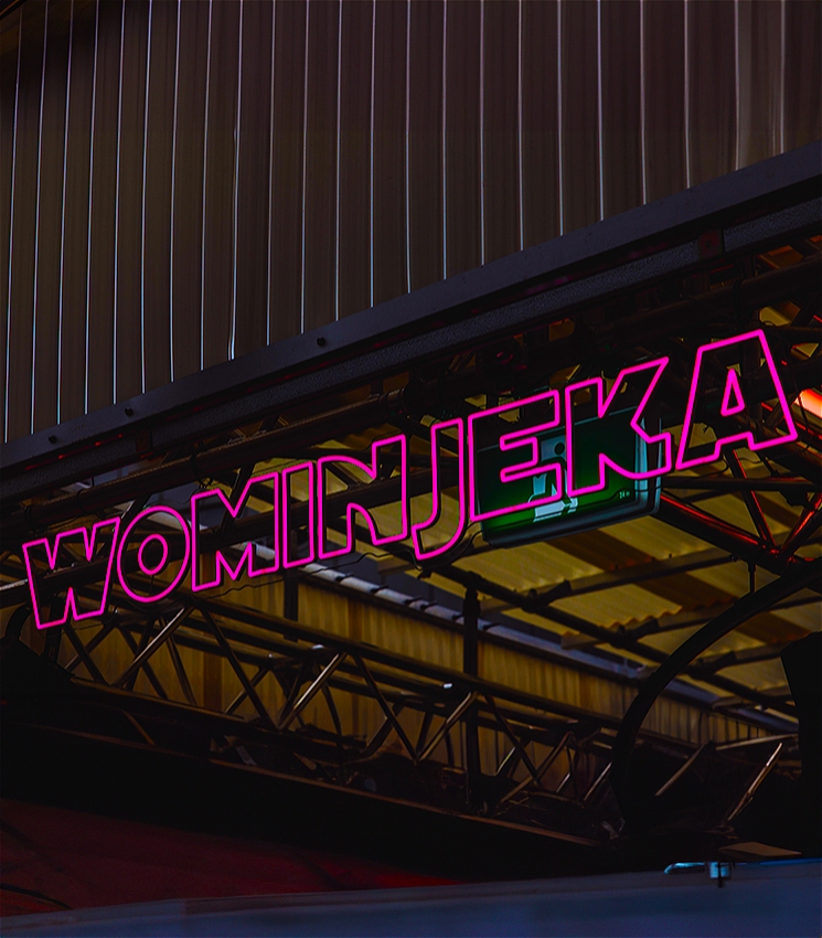 A sign outlined in pink that reads: Wominjeka. Behind the sign comprising of roof panels and railings.