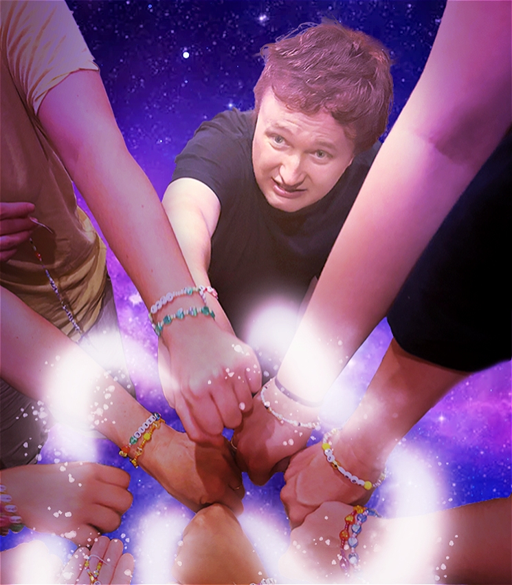 The image is a photo that captures lots of hands coming to the centre of the image, showing off their friendship bracelets. The bracelets are a collection beads - some of which have letters, some plane colours and some sparkle. We also see Ben, a masculine white person with short brown hair, wearing a black t-shirt and pants who is trying to join in with the group with his hand. His face shows a distressed expression.