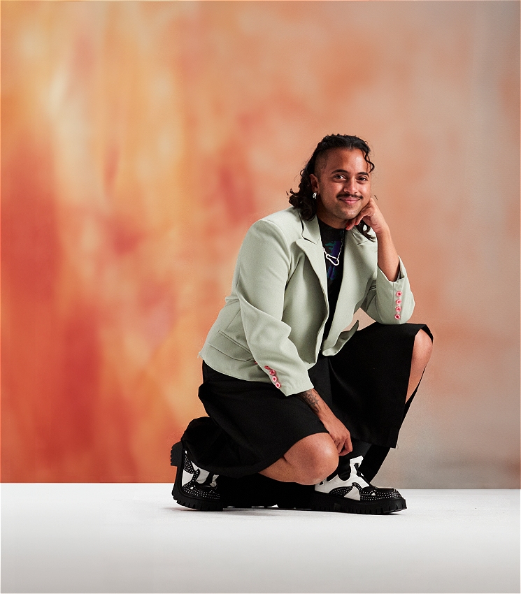 Krishna, a Brown trans person in their 30s, wears a green blazer and black skirt. Krishna is posed bent down on one knee, looking straight at the camera with a friendly expression.
