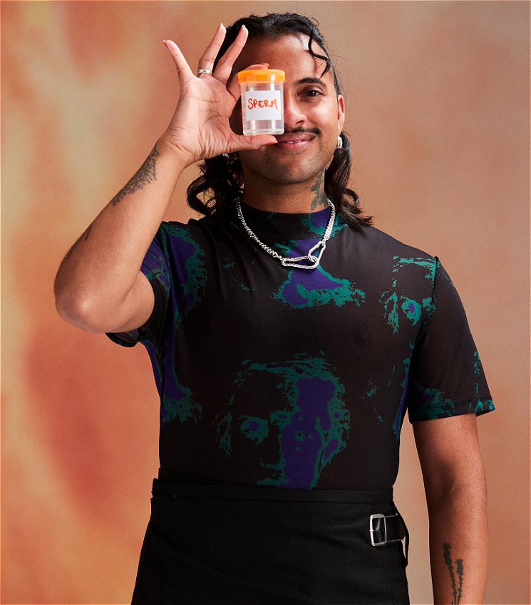 Krishna, a Brown trans person in their 30s, wears a dark black shirt with splotches of blue and green. They hold a sample collection vial in front of their face labelled "sperm". They look straight at the camera with a friendly expression.