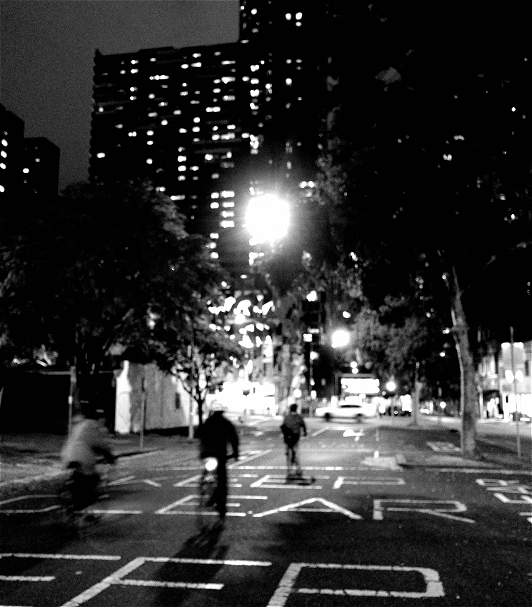Black and white photograph, blurry picture of 3 cyclists riding up a city street (view from behind the cyclists).